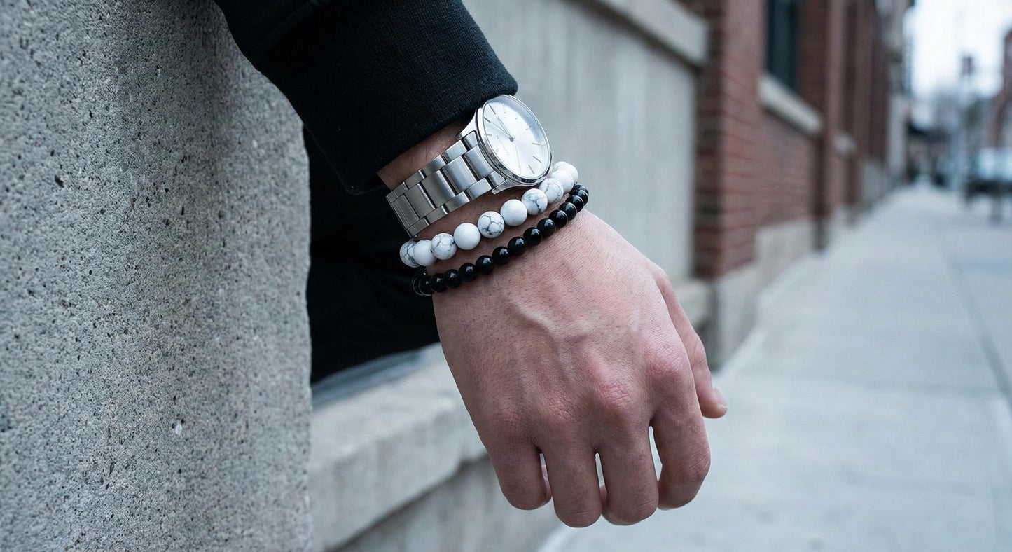 Male wrist wearing silver watch and black and white beaded bracelets against urban sidewalk background