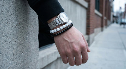 Male wrist wearing silver watch and black and white beaded bracelets against urban sidewalk background