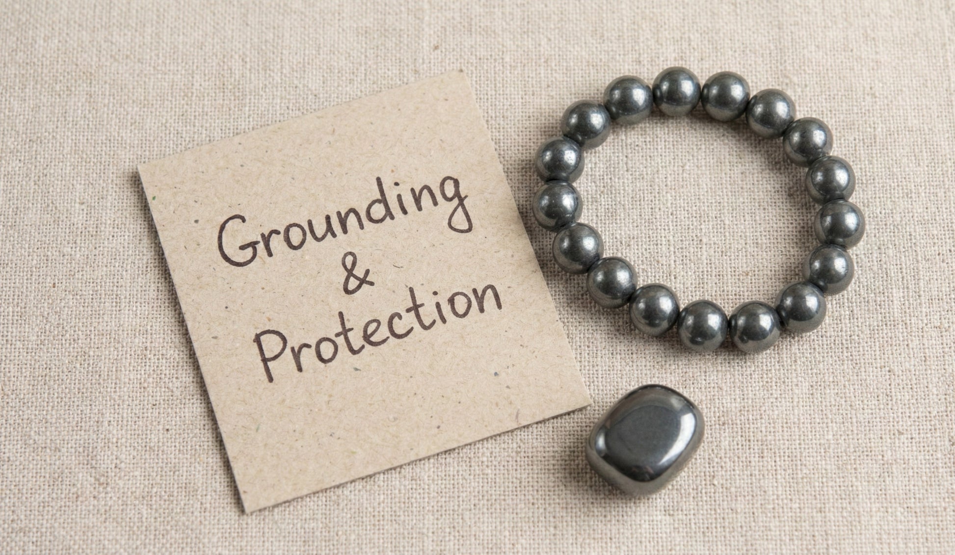 Hematite bracelet and stone next to a paper note reading Grounding & Protection on beige fabric