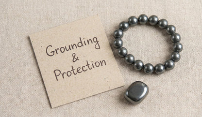 Hematite bracelet and stone next to a paper note reading Grounding & Protection on beige fabric