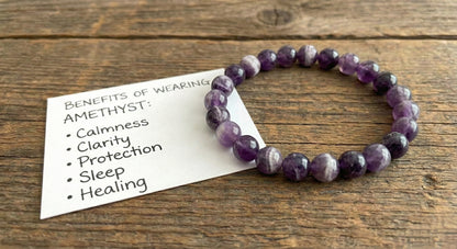 Amethyst beaded bracelet on wood surface with card listing benefits: calmness, clarity, protection, sleep, healing