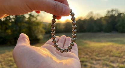 Hand holding reflective metallic bead bracelet outdoors at sunset with blurred trees and field background