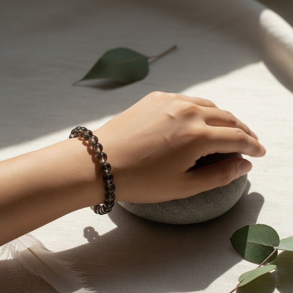 Hand wearing a smoky quartz bead bracelet resting on a smooth stone with eucalyptus leaves and soft light
