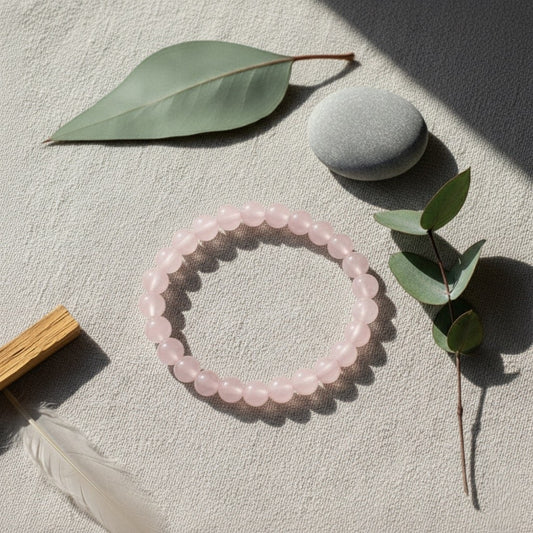 Pink rose quartz beaded bracelet with eucalyptus leaves, smooth gray stone, and wooden incense holder on textured fabric